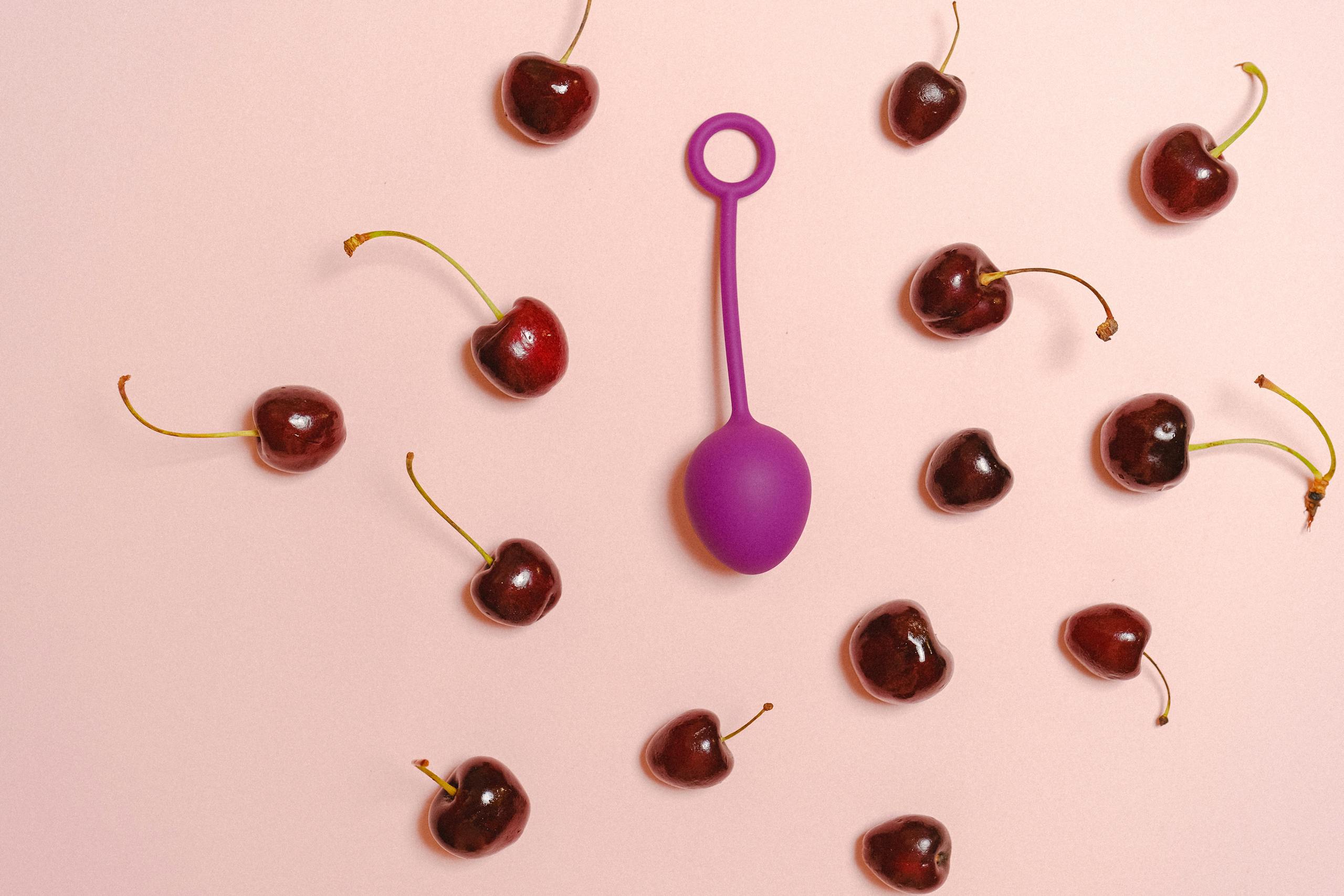 Vibrant still life of a purple silicone toy surrounded by cherries on a pastel pink background.